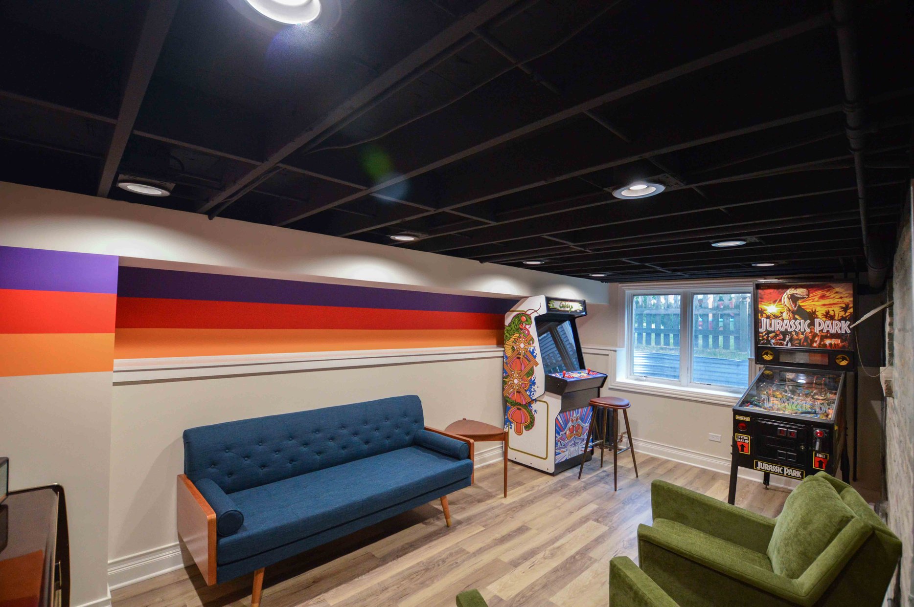 Blue couch, Green chairs, and two arcade games in a finished basement. Bold striped wall mural. 