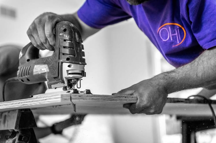 man with electric saw sawing a piece of wood wearing a purple shirt with OHi logo 