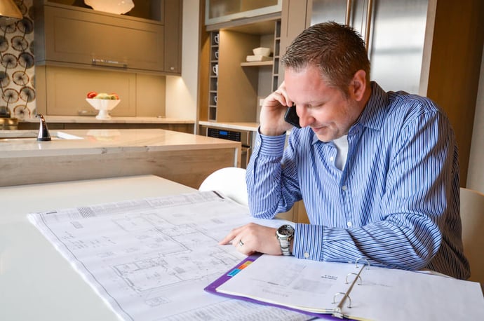 Man in blue button up on the phone sitting at a counter with plans and a folder 