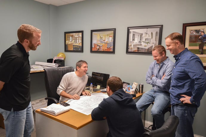 Five men sitting in an office around a desk laughing 