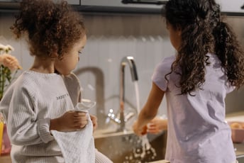 Two kids standing at the sink doing dishes together in a kitchen as the faucer is running.